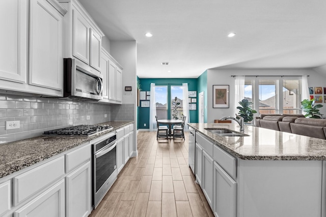 kitchen featuring a sink, an island with sink, stainless steel appliances, backsplash, and light wood-style flooring