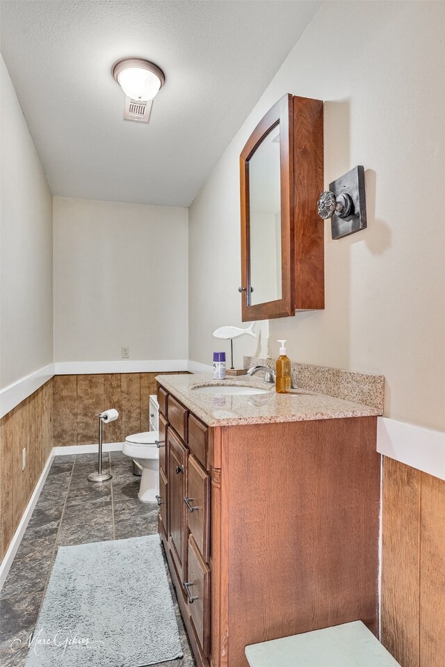 half bath featuring vanity, stone finish flooring, visible vents, toilet, and wainscoting