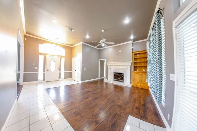 unfurnished living room with crown molding, ceiling fan, visible vents, a tiled fireplace, and tile patterned floors