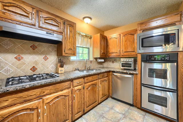 kitchen with a sink, appliances with stainless steel finishes, a textured ceiling, brown cabinetry, and backsplash