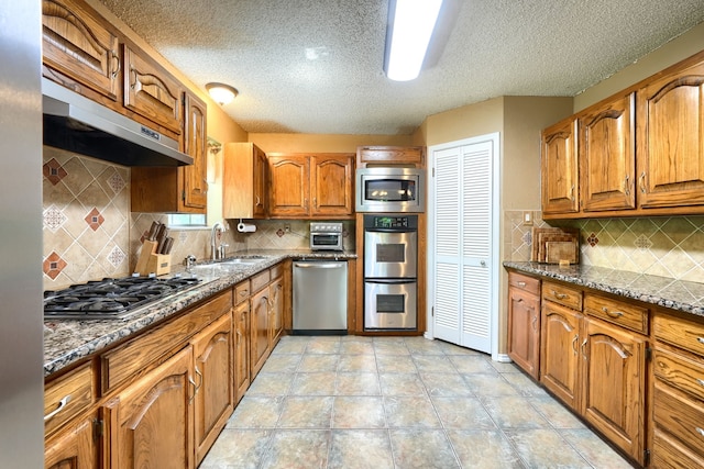 kitchen with a sink, appliances with stainless steel finishes, a textured ceiling, under cabinet range hood, and brown cabinets