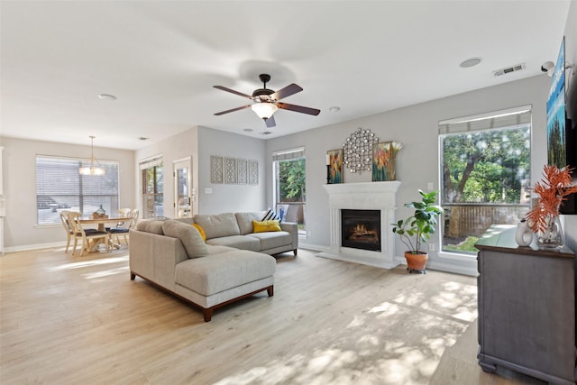 living room featuring visible vents, baseboards, a warm lit fireplace, light wood-type flooring, and ceiling fan with notable chandelier