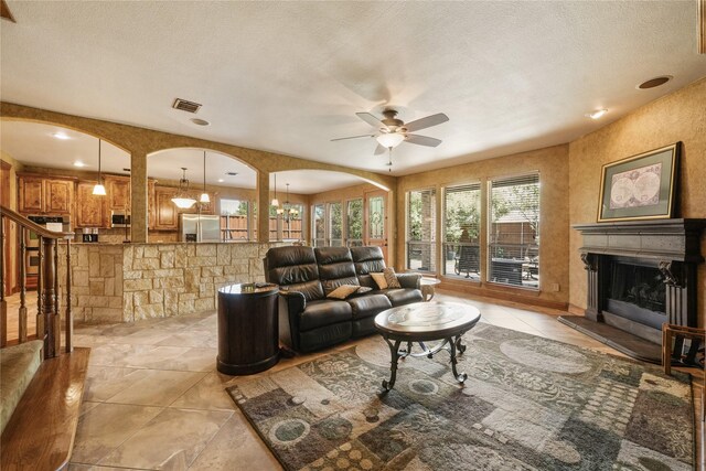 living area with light tile patterned floors, visible vents, a fireplace with raised hearth, arched walkways, and ceiling fan with notable chandelier
