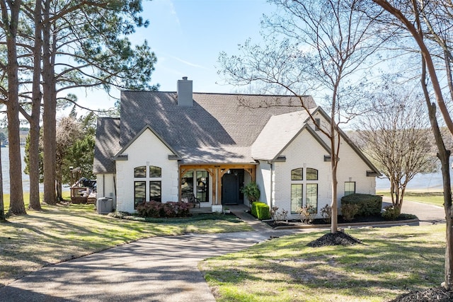 french provincial home with roof with shingles, a front yard, brick siding, a chimney, and central AC unit