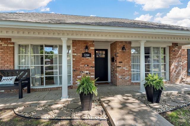doorway to property featuring brick siding, a shingled roof, and covered porch