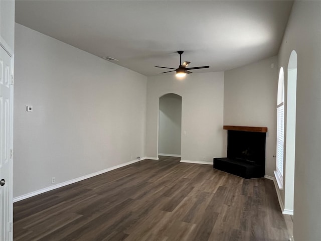 unfurnished living room featuring a ceiling fan, a fireplace with raised hearth, visible vents, dark wood finished floors, and arched walkways