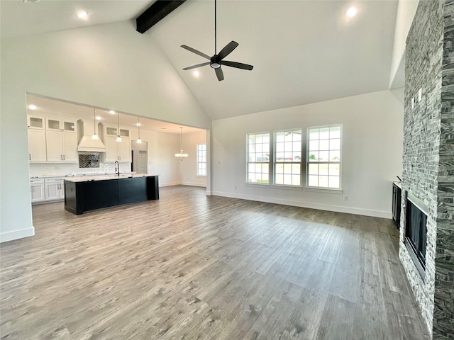 unfurnished living room featuring a sink, beam ceiling, a fireplace, high vaulted ceiling, and ceiling fan with notable chandelier