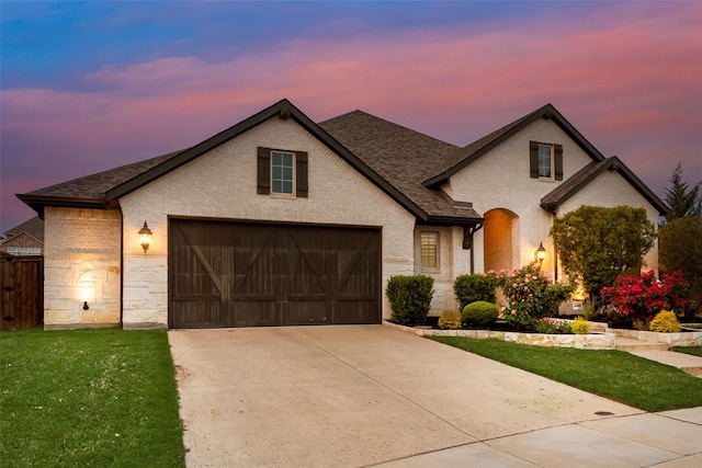 french country inspired facade with a lawn, an attached garage, driveway, and roof with shingles