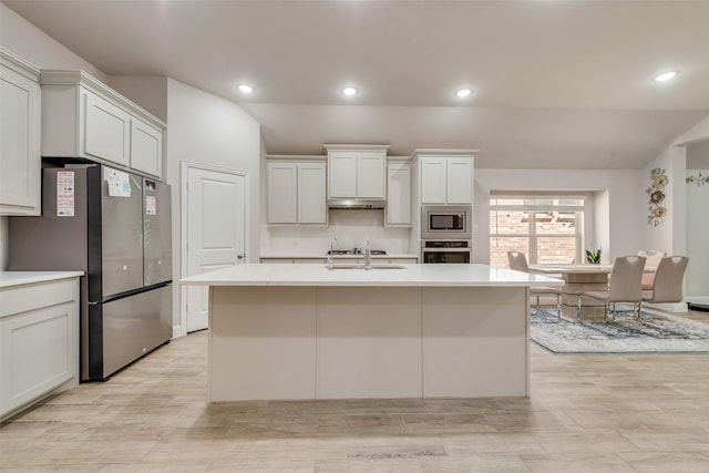 kitchen with light countertops, under cabinet range hood, a kitchen island with sink, appliances with stainless steel finishes, and a sink