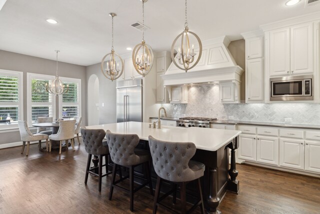 kitchen with custom exhaust hood, built in appliances, an inviting chandelier, a center island with sink, and visible vents