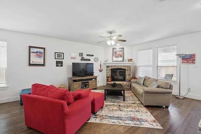 living area with a stone fireplace, baseboards, dark wood-type flooring, and ceiling fan