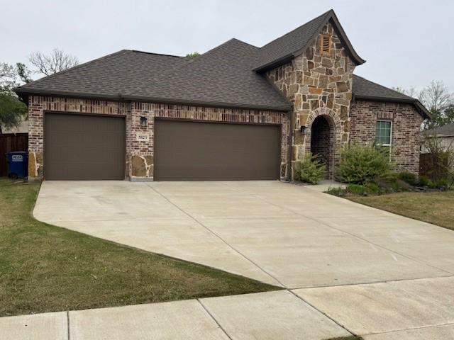 french country home with roof with shingles, driveway, stone siding, an attached garage, and brick siding