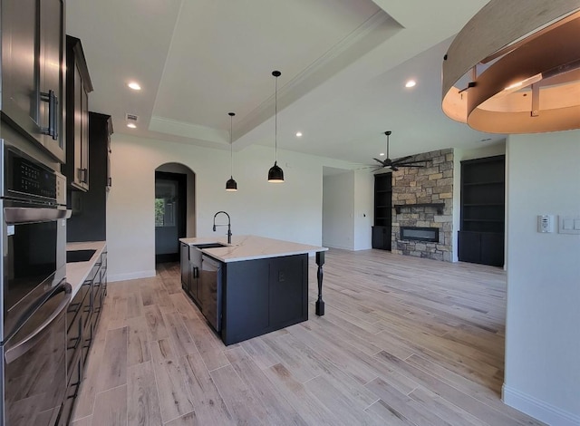 kitchen featuring a ceiling fan, a center island with sink, a tray ceiling, stainless steel oven, and arched walkways