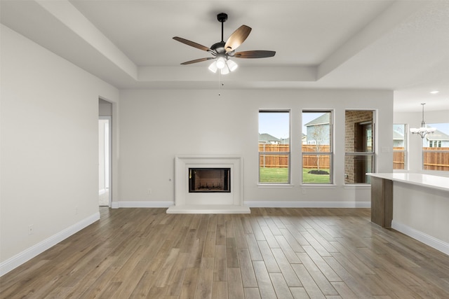 unfurnished living room with baseboards, a raised ceiling, a fireplace with raised hearth, wood finished floors, and ceiling fan with notable chandelier