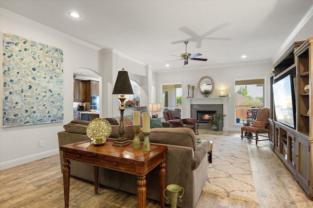living area featuring arched walkways, a brick fireplace, a ceiling fan, crown molding, and light wood finished floors