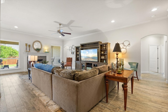 living room featuring crown molding, light wood-style flooring, arched walkways, a ceiling fan, and a brick fireplace