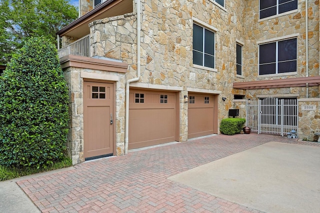 exterior space with an attached garage, a balcony, stone siding, and decorative driveway