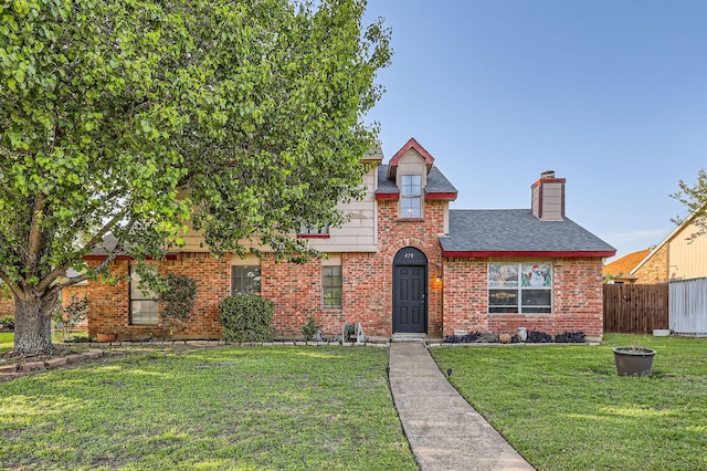 tudor house with roof with shingles, a chimney, a front yard, brick siding, and fence