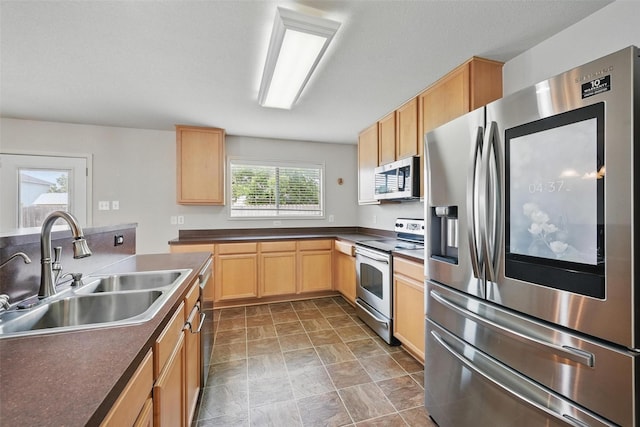 kitchen with dark countertops, appliances with stainless steel finishes, a sink, light brown cabinets, and stone finish flooring