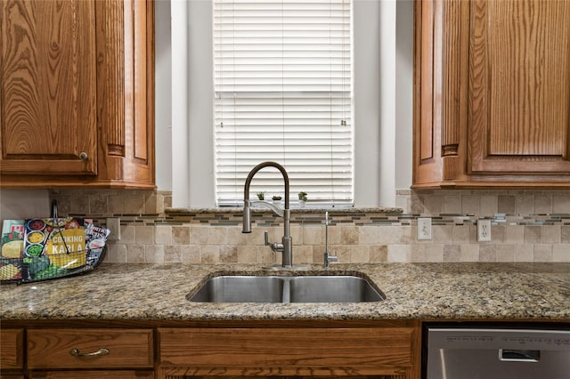kitchen with brown cabinetry, backsplash, light stone countertops, dishwasher, and a sink