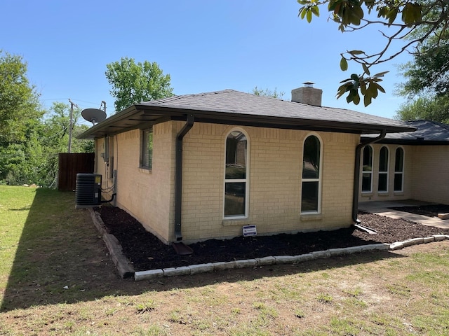 view of property exterior featuring roof with shingles, a chimney, brick siding, central AC, and a yard