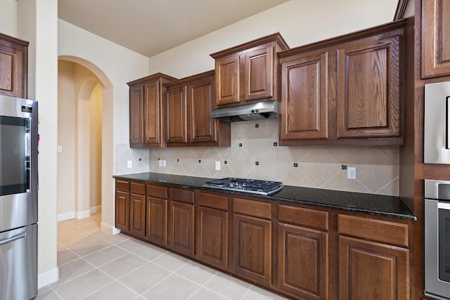 kitchen featuring dark stone countertops, light tile patterned floors, under cabinet range hood, stainless steel appliances, and arched walkways