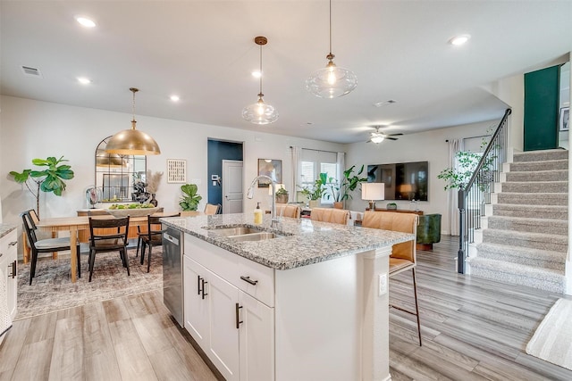 kitchen with light wood-type flooring, an island with sink, light stone countertops, stainless steel dishwasher, and a sink