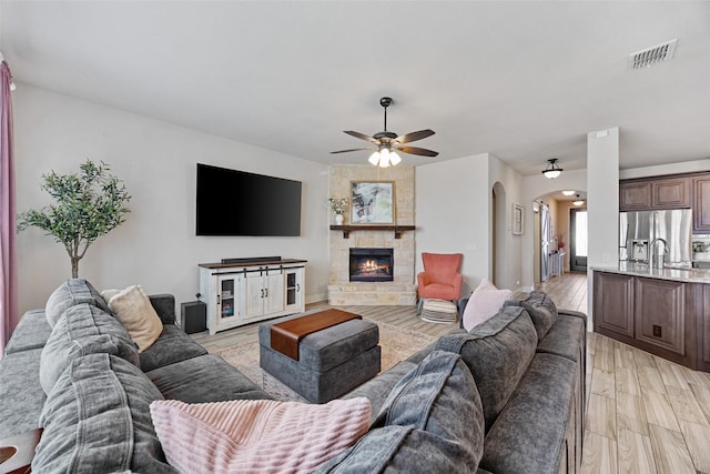 living room featuring visible vents, arched walkways, a stone fireplace, light wood-type flooring, and a ceiling fan
