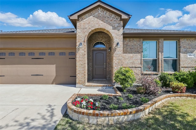 doorway to property featuring an attached garage, roof with shingles, concrete driveway, and brick siding