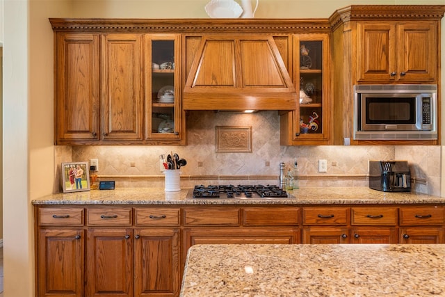 kitchen with brown cabinetry, custom exhaust hood, light stone countertops, and stainless steel appliances