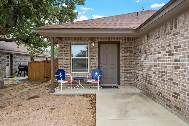entrance to property with brick siding, fence, a patio, and a shingled roof