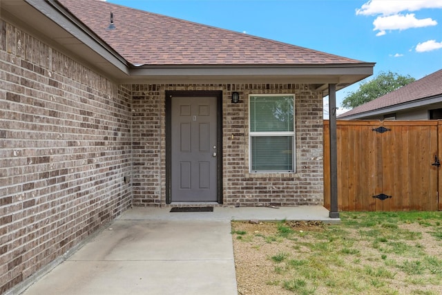 view of exterior entry with brick siding, roof with shingles, and fence