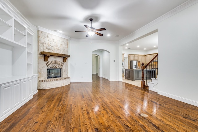 unfurnished living room featuring crown molding, a stone fireplace, wood finished floors, ceiling fan, and arched walkways
