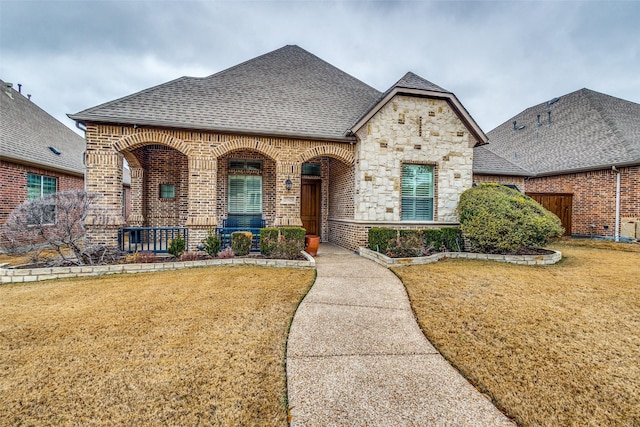 french country home featuring stone siding, fence, a front yard, brick siding, and roof with shingles