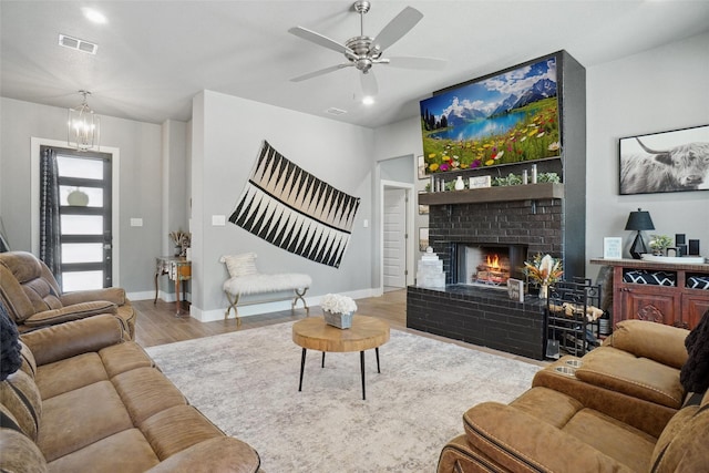 living room with wood finished floors, ceiling fan with notable chandelier, baseboards, a brick fireplace, and visible vents