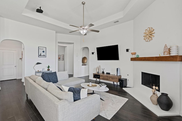 living room featuring wood finished floors, a tray ceiling, arched walkways, and a fireplace with raised hearth