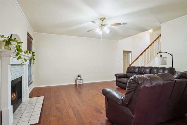 living area featuring a tiled fireplace, wood finished floors, stairs, ceiling fan, and crown molding