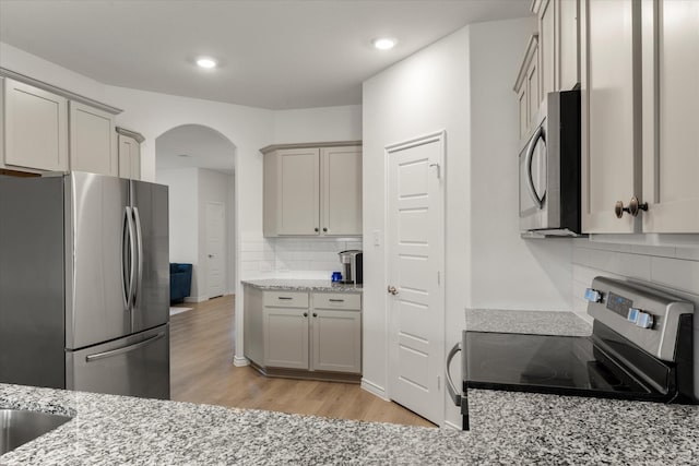 kitchen featuring stainless steel appliances, arched walkways, light wood-type flooring, light stone countertops, and tasteful backsplash