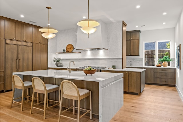 kitchen with stainless steel gas stovetop, a kitchen island with sink, custom exhaust hood, and light wood-type flooring