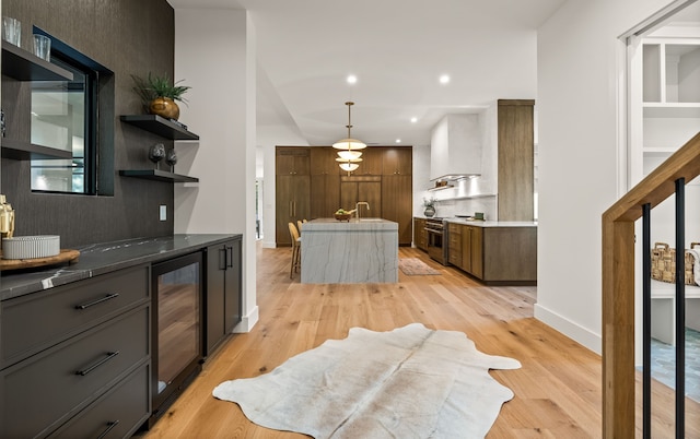 kitchen with stainless steel range, a kitchen island with sink, light wood finished floors, wall chimney exhaust hood, and wine cooler