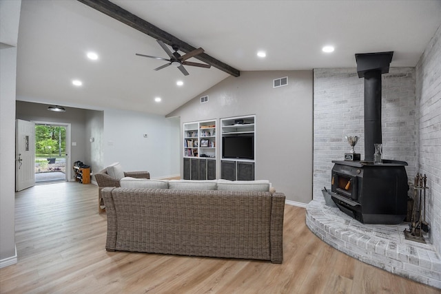 living area with a ceiling fan, vaulted ceiling with beams, a wood stove, light wood finished floors, and visible vents