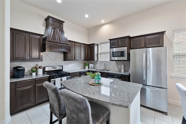 kitchen with stainless steel appliances, a center island, dark brown cabinets, light stone countertops, and premium range hood
