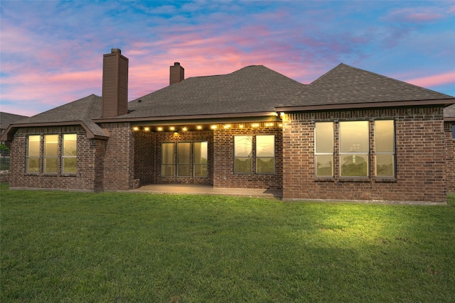 back of property at dusk featuring a shingled roof, brick siding, a chimney, and a lawn