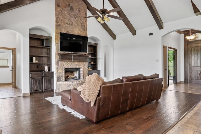 living room featuring beam ceiling, high vaulted ceiling, ceiling fan, arched walkways, and a stone fireplace
