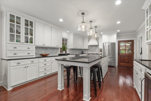 kitchen with high end fridge, dark countertops, custom exhaust hood, a kitchen bar, and ornamental molding