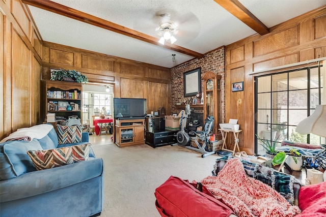 carpeted living room with a ceiling fan, a textured ceiling, wooden walls, a brick fireplace, and beam ceiling