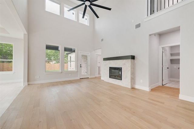 unfurnished living room with a towering ceiling, light wood finished floors, a ceiling fan, a healthy amount of sunlight, and a high end fireplace