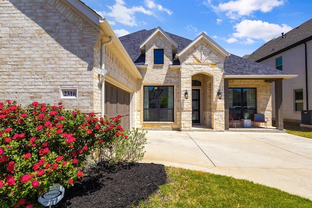 view of front of property with central AC, stone siding, concrete driveway, a garage, and a shingled roof