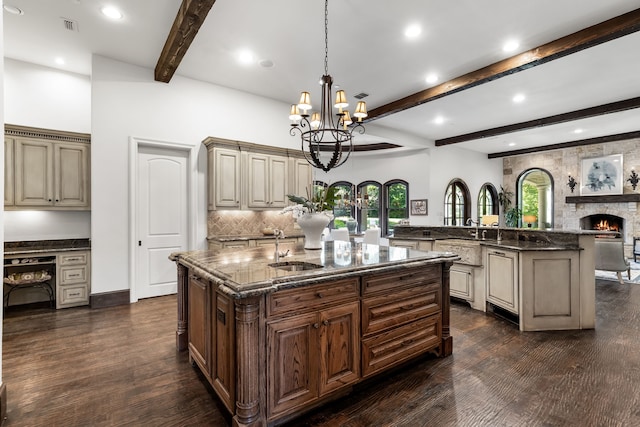kitchen featuring beam ceiling, a sink, dark stone counters, a center island with sink, and a chandelier