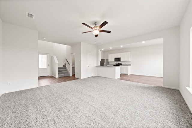 unfurnished living room featuring carpet, visible vents, a ceiling fan, and stairs